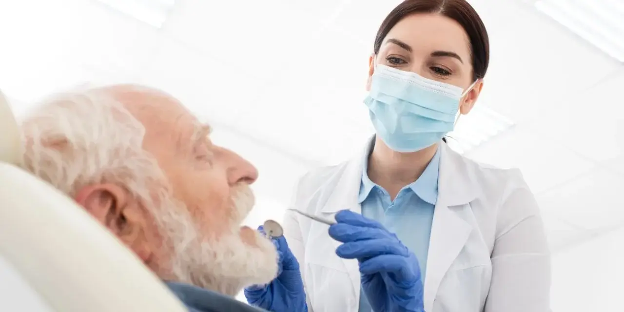 Dentystka w masce bada zęby starszego mężczyzny przy użyciu narzędzi stomatologicznych.