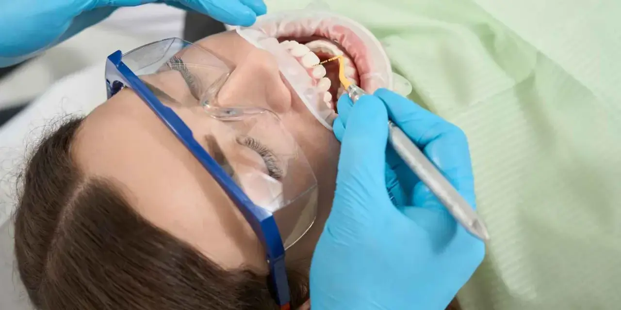Pacjentka w okularach ochronnych poddawana zabiegowi higienizacji. Dentysta w niebieskich rękawiczkach używa narzędzia do czyszczenia zębów.