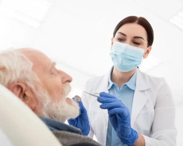 Dentystka w masce bada zęby starszego mężczyzny przy użyciu narzędzi stomatologicznych.