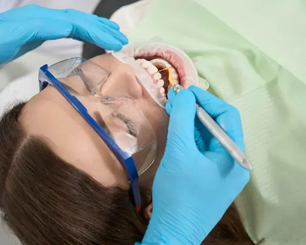Pacjentka w okularach ochronnych poddawana zabiegowi higienizacji. Dentysta w niebieskich rękawiczkach używa narzędzia do czyszczenia zębów.
