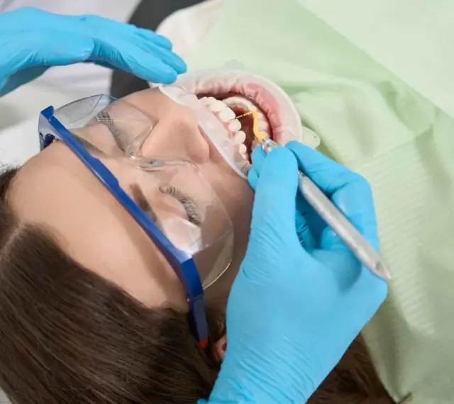 Pacjentka w okularach ochronnych poddawana zabiegowi higienizacji. Dentysta w niebieskich rękawiczkach używa narzędzia do czyszczenia zębów.