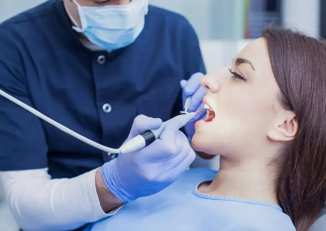 Dentysta w rękawiczkach wykonuje zabieg. Pacjentka ma otwarte usta. Zastanawiasz się, ile kosztuje znieczulenie u dentysty?