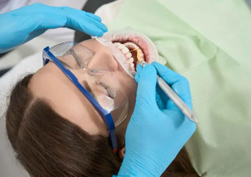 Pacjentka w okularach ochronnych poddawana zabiegowi higienizacji. Dentysta w niebieskich rękawiczkach używa narzędzia do czyszczenia zębów.